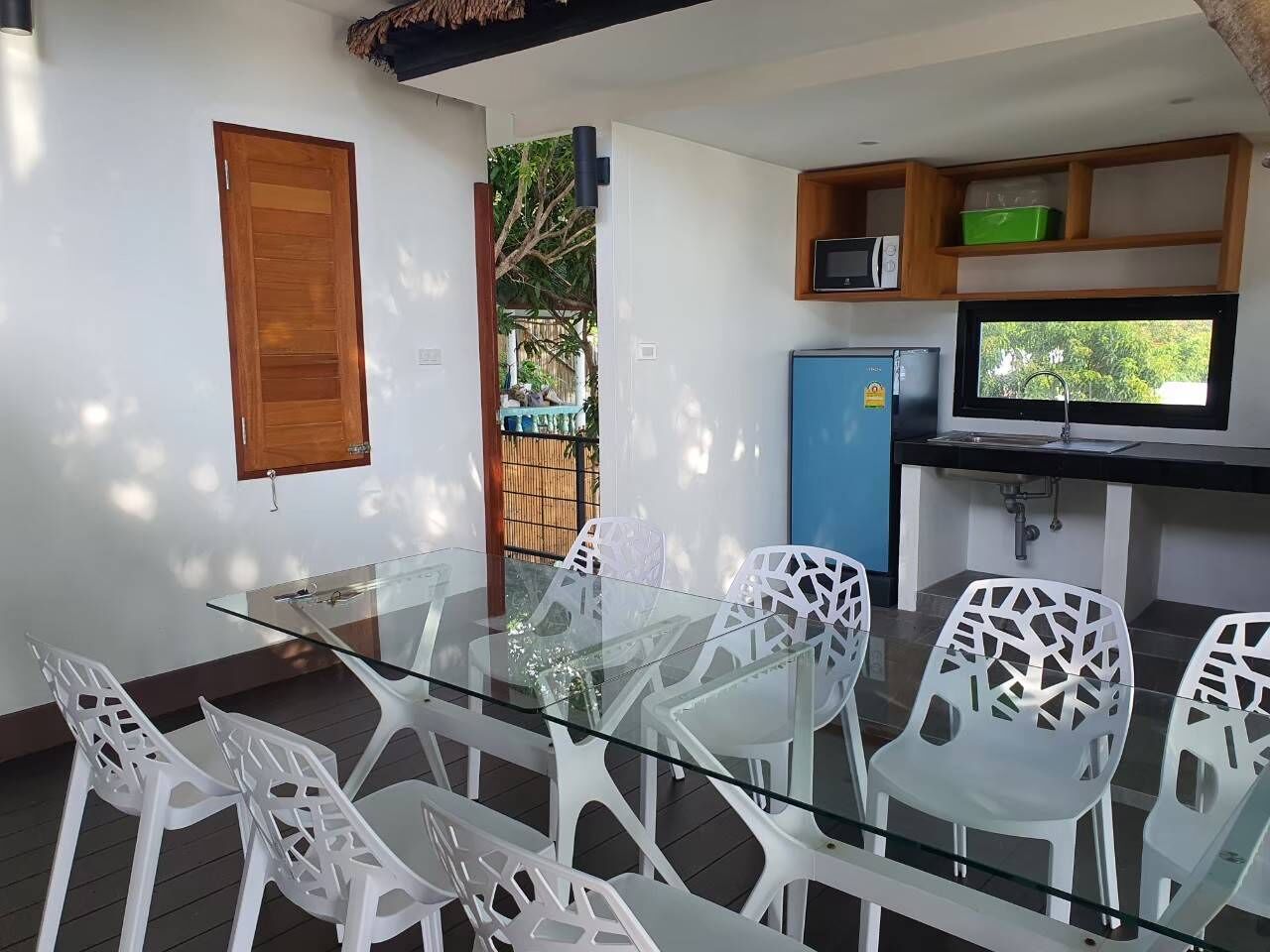 A glass-top dining table with white chairs, a kitchen area with a microwave and a refrigerator, and a wooden door.