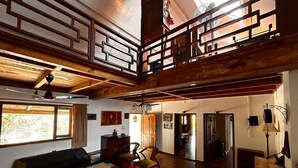 A spacious living area with a high ceiling, wooden beams, a sofa, chairs, a dining table, and a view through a window. - Emerald Trail (Nainital)