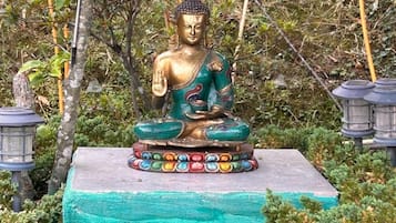 A golden Buddha statue on a pedestal with a colorful base, surrounded by greenery and a stone wall.