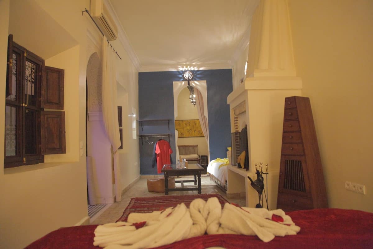 Un pasillo estrecho conduce a un dormitorio con cama, mesa y silla. Hay una toalla roja y blanca sobre la cama, un armario de madera y un espejo en la pared.