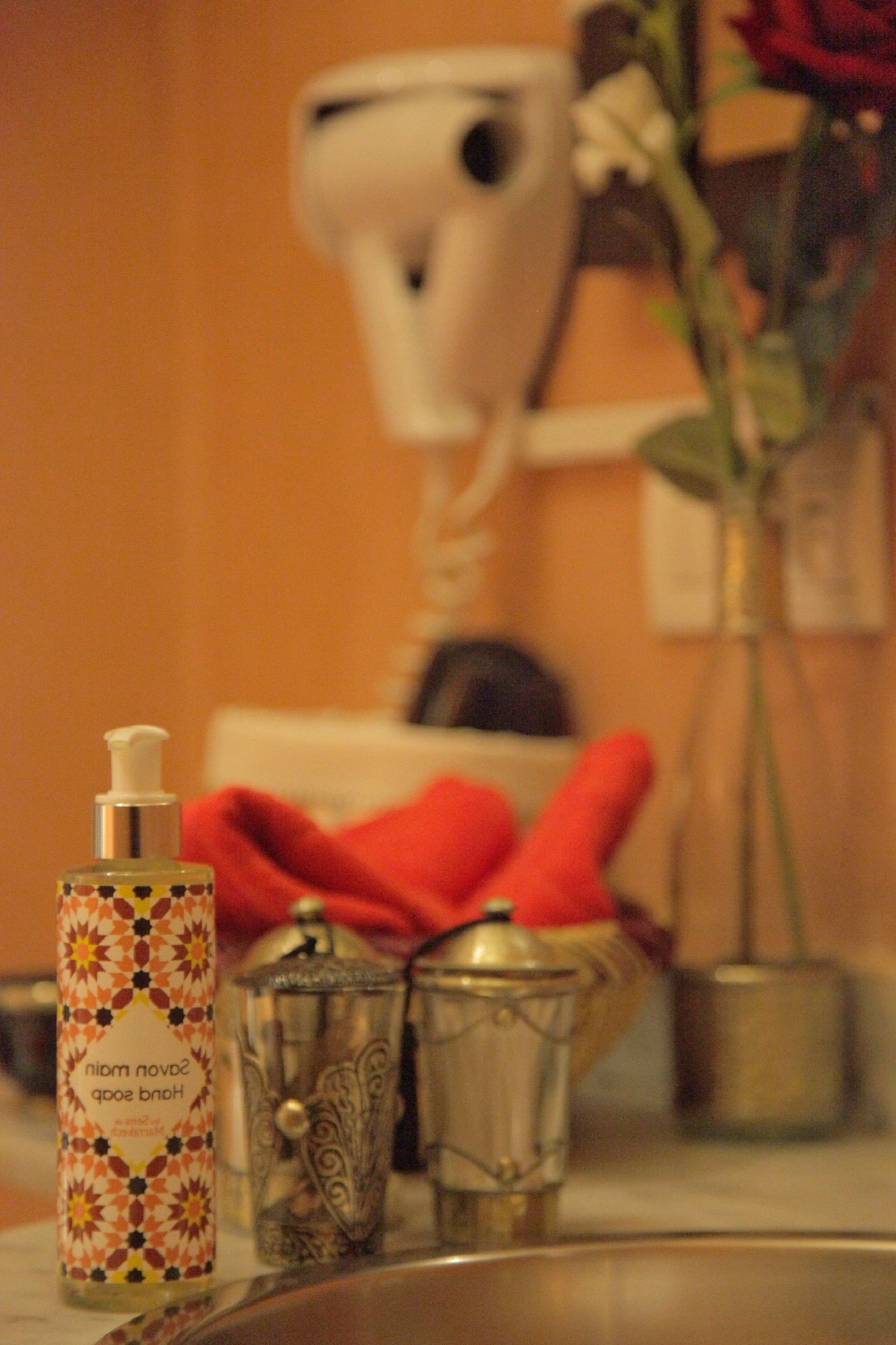 A bathroom with a hairdryer, a bottle of hand soap, a glass container, and a vase with a flower.