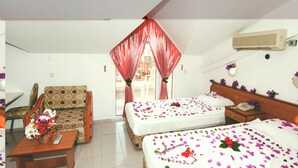 Une chambre d’hôtel avec deux lits, un canapé, une petite table sur laquelle est posé un vase de fleurs, et une fenêtre avec des rideaux rouges.