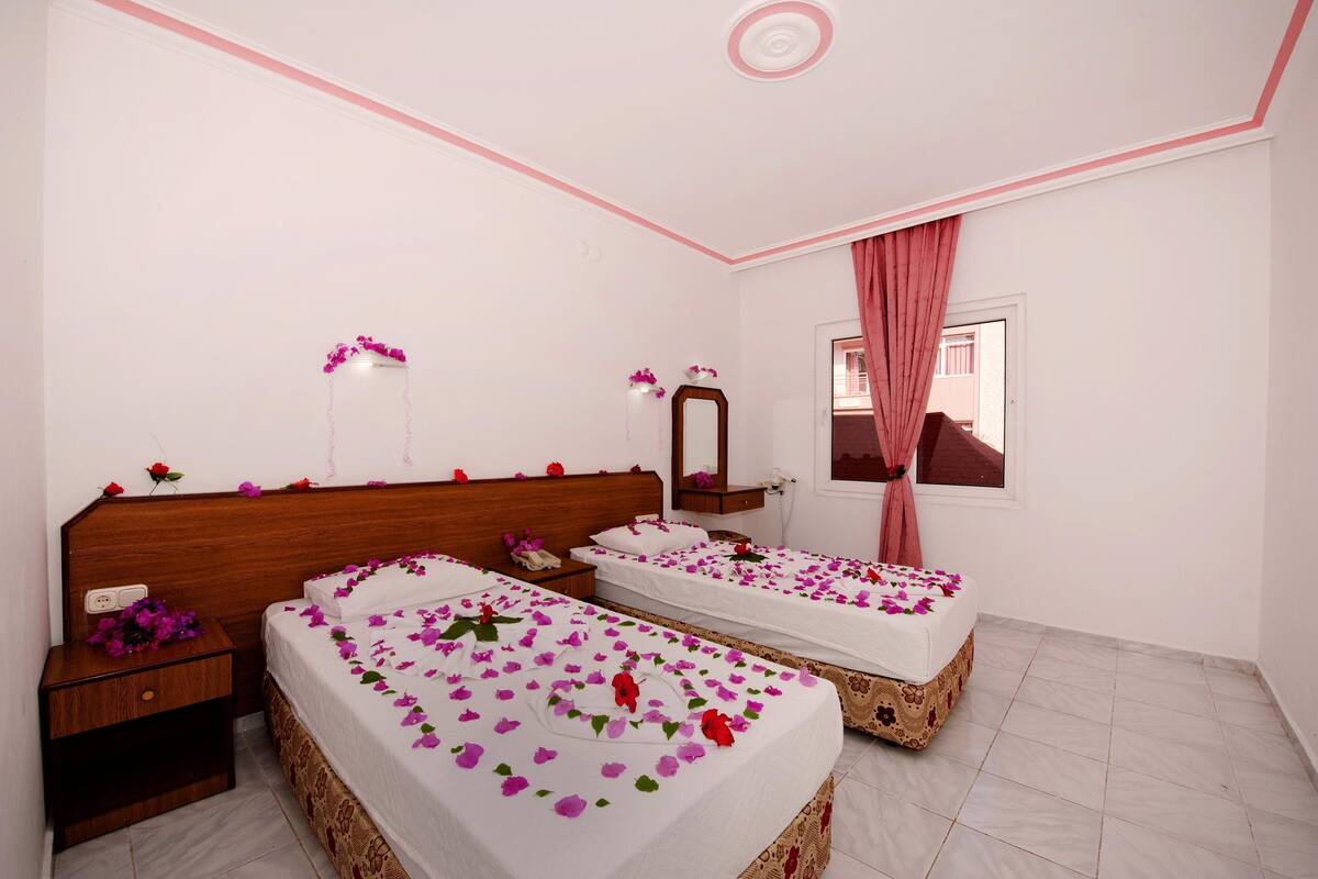 Une chambre d’hôtel avec deux lits, une tête de lit en bois, une table de chevet, un miroir et une fenêtre avec des rideaux roses.