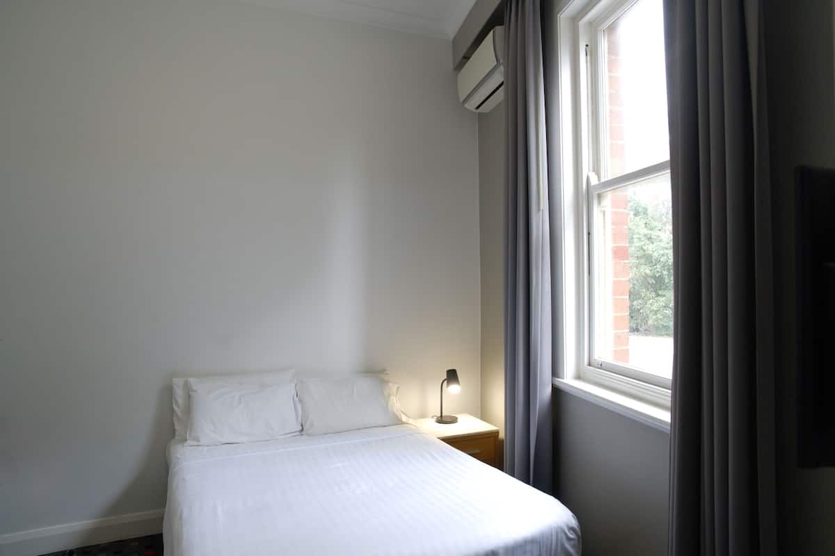 Una habitación de hotel con cama, mesita de noche, lámpara y ventana con cortinas.