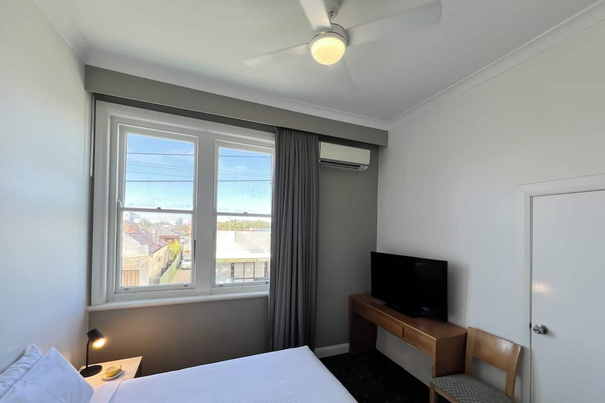 Habitación de hotel con cama, escritorio con televisor, silla, ventana con cortinas y unidad de aire acondicionado.