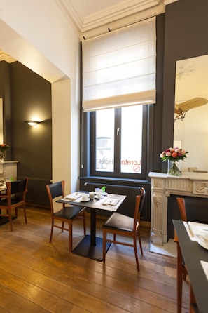 A dining area with a table set for two, a fireplace, and a window with a view of the street. - Hotel Loreto (Bruges)