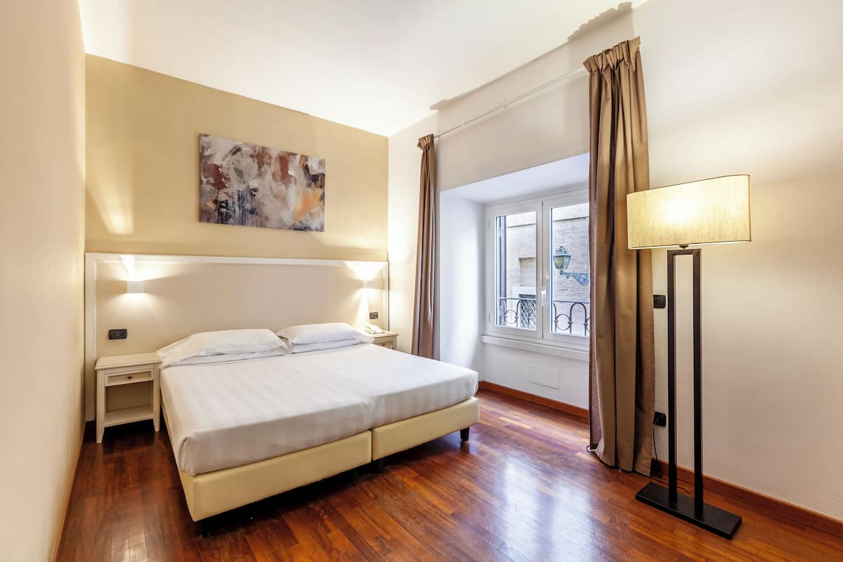 A bedroom with a bed, bedside lamps, a painting on the wall, and a window with curtains.