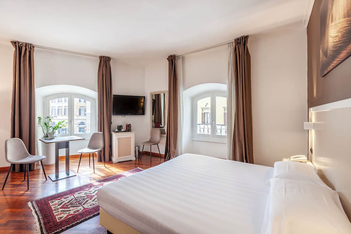 A hotel room with a large bed, a desk with two chairs, a TV, and a view of buildings through arched windows.