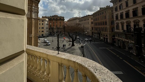 Un balcó amb vista a un carrer de la ciutat, edificis i un cel amb núvols dispersos.