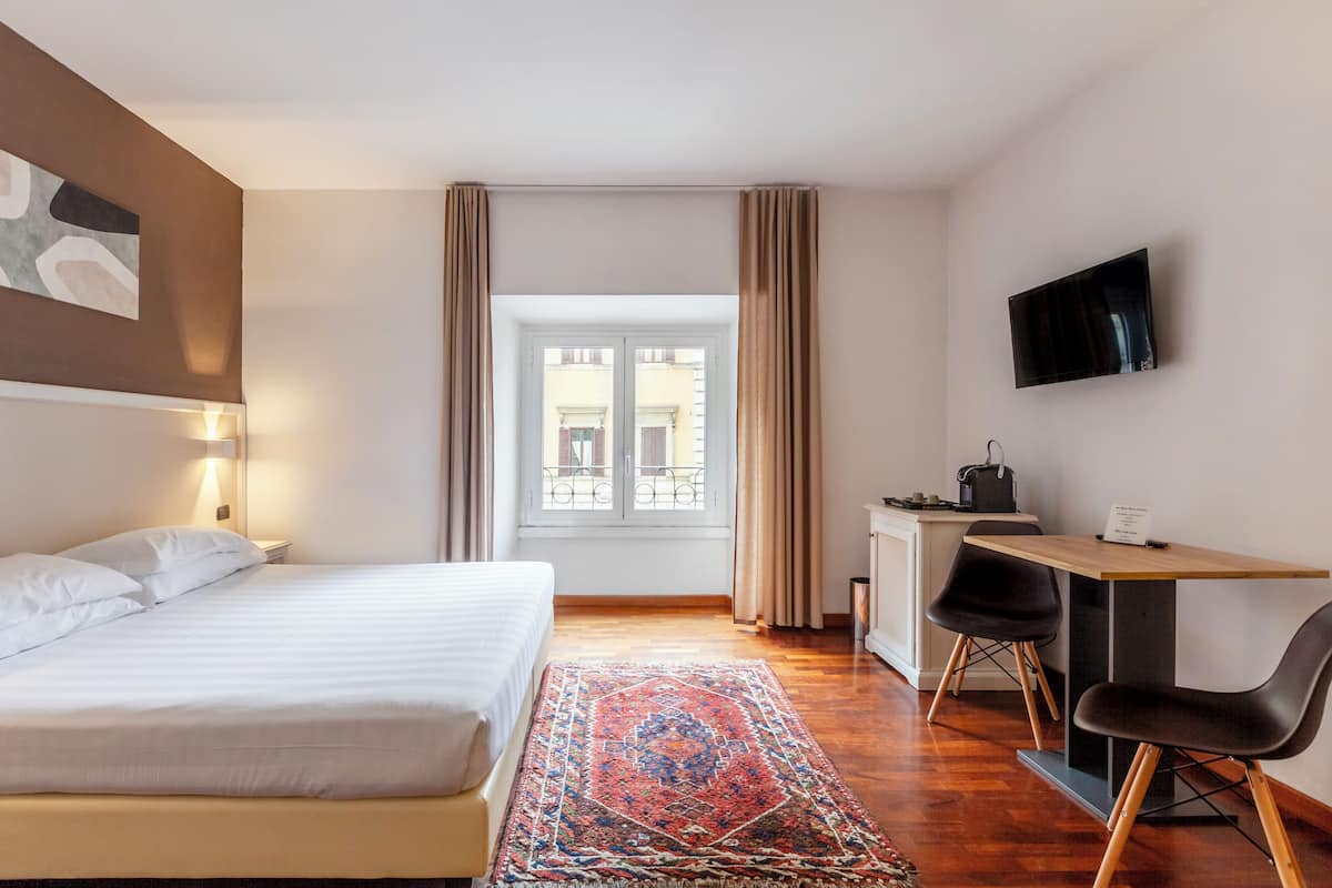 A hotel room with a bed, desk, chairs, a TV, and a window with curtains.
