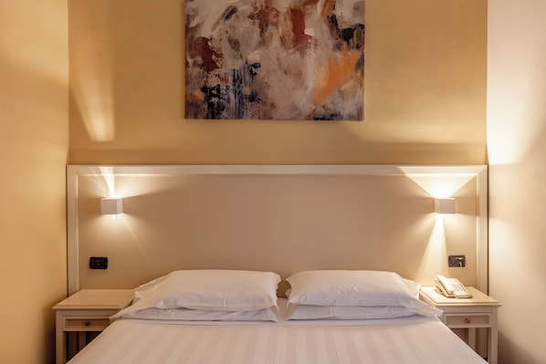 Una cama bien hecha con sábanas y almohadas blancas, una mesita de noche con lámpara y un cuadro abstracto colgado en la pared.