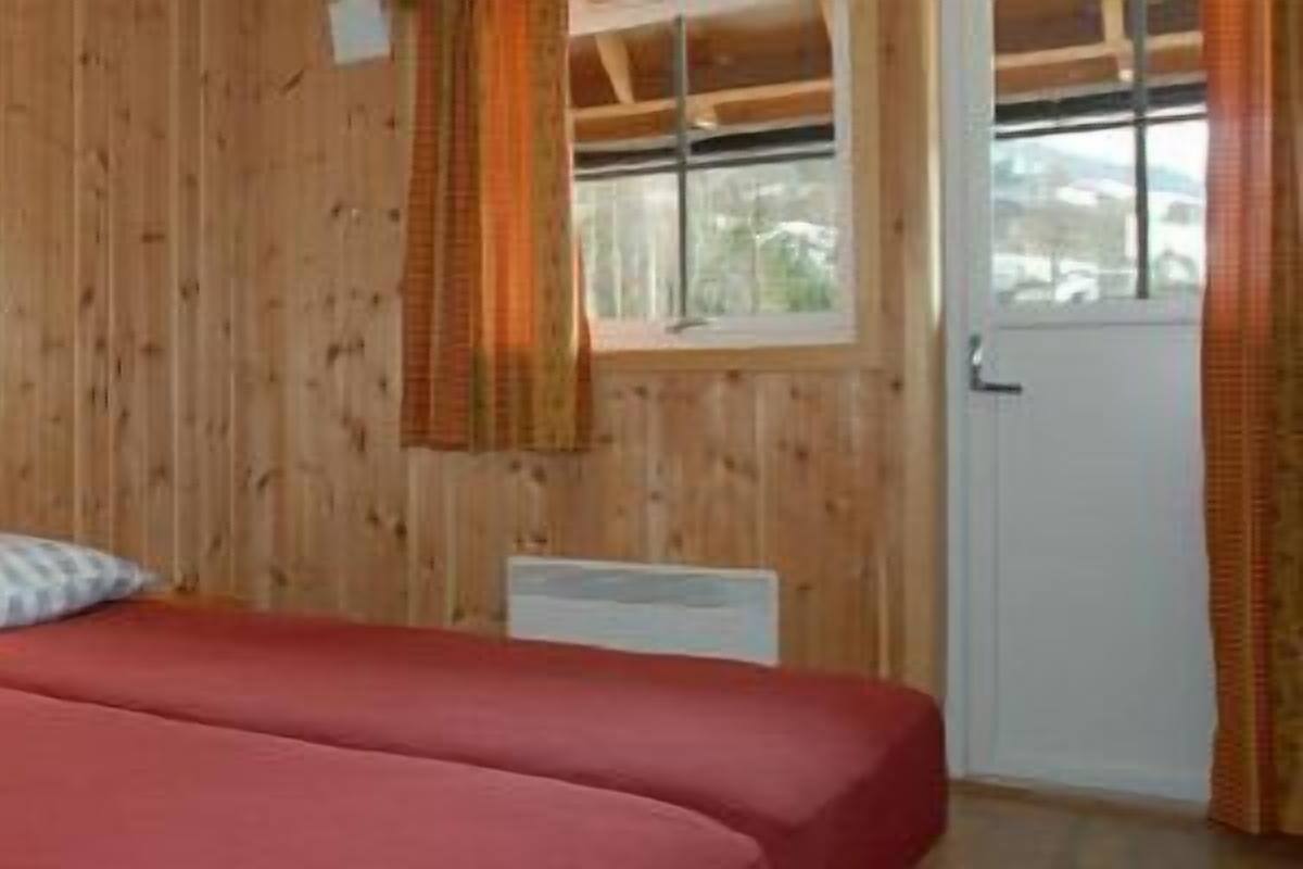 Huone, jonka seinät ovat puuta, sängyssä on kaksi tyynyä, ikkunassa on verhot ja huoneessa on ovi.