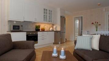A modern kitchen with white cabinets, a microwave, and a refrigerator. There are two brown sofas and a wooden coffee table with candles.