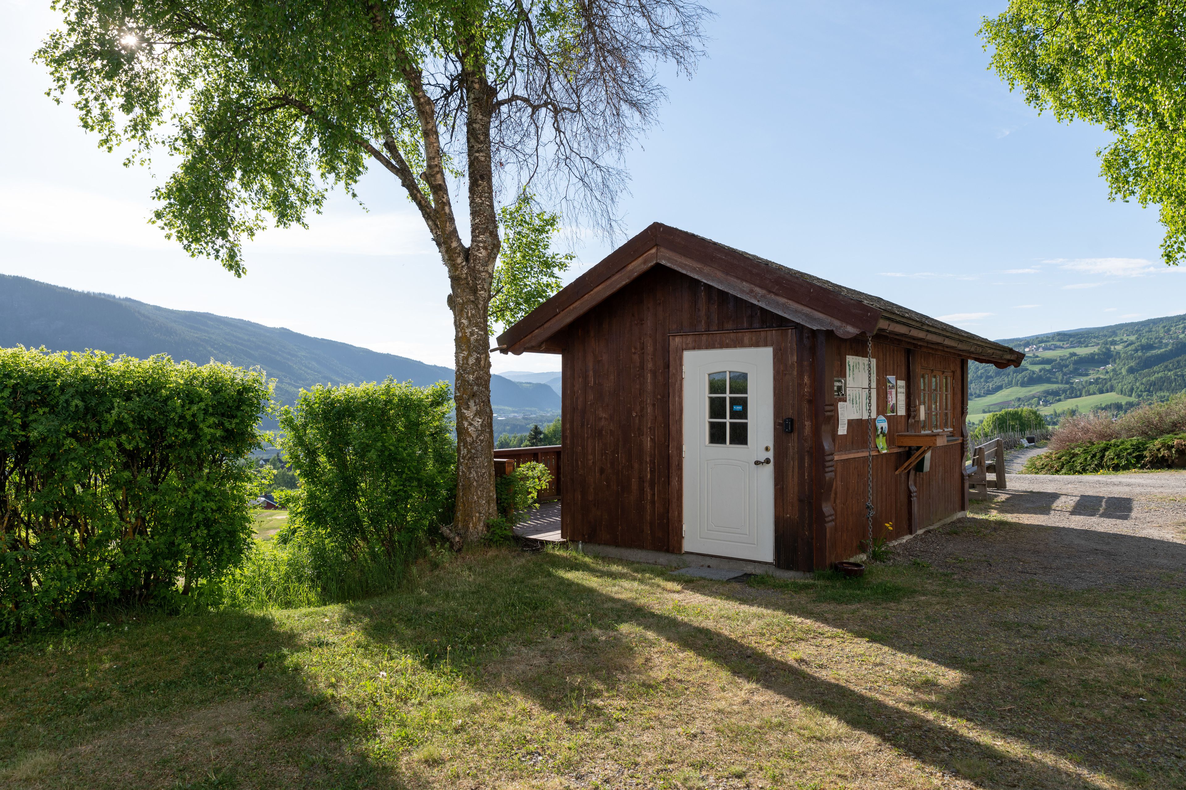 A wooden cabin with a white door, surrounded by greenery and hills in the background.