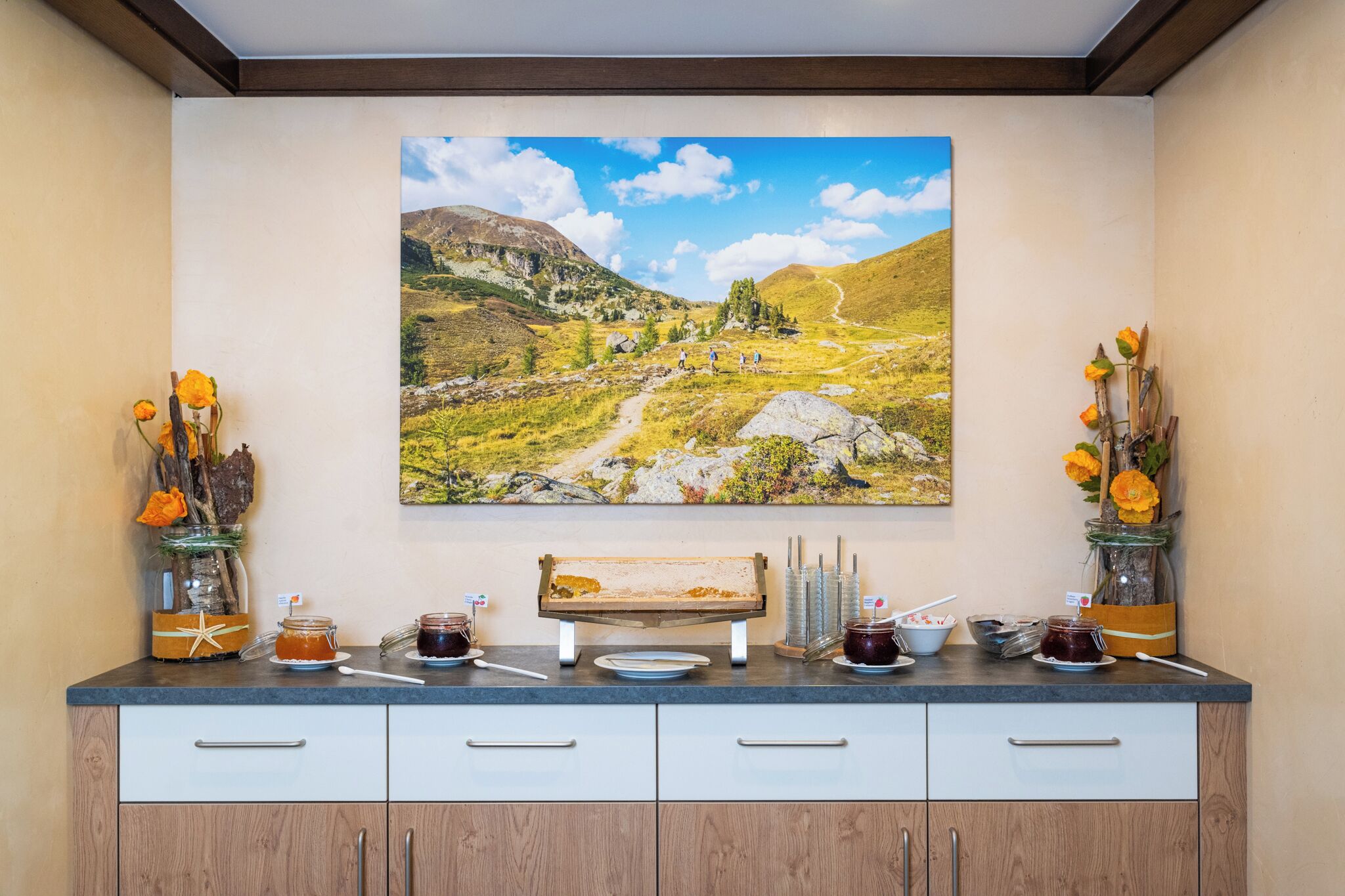 A kitchen counter with a painting of a landscape, jars of jam, and a bench.