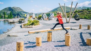 A person balancing on a log over a gravel area with wooden posts, with mountains and a body of water in the background. - Ski- & Wanderhotel Berghof (Bad Kleinkirchheim)