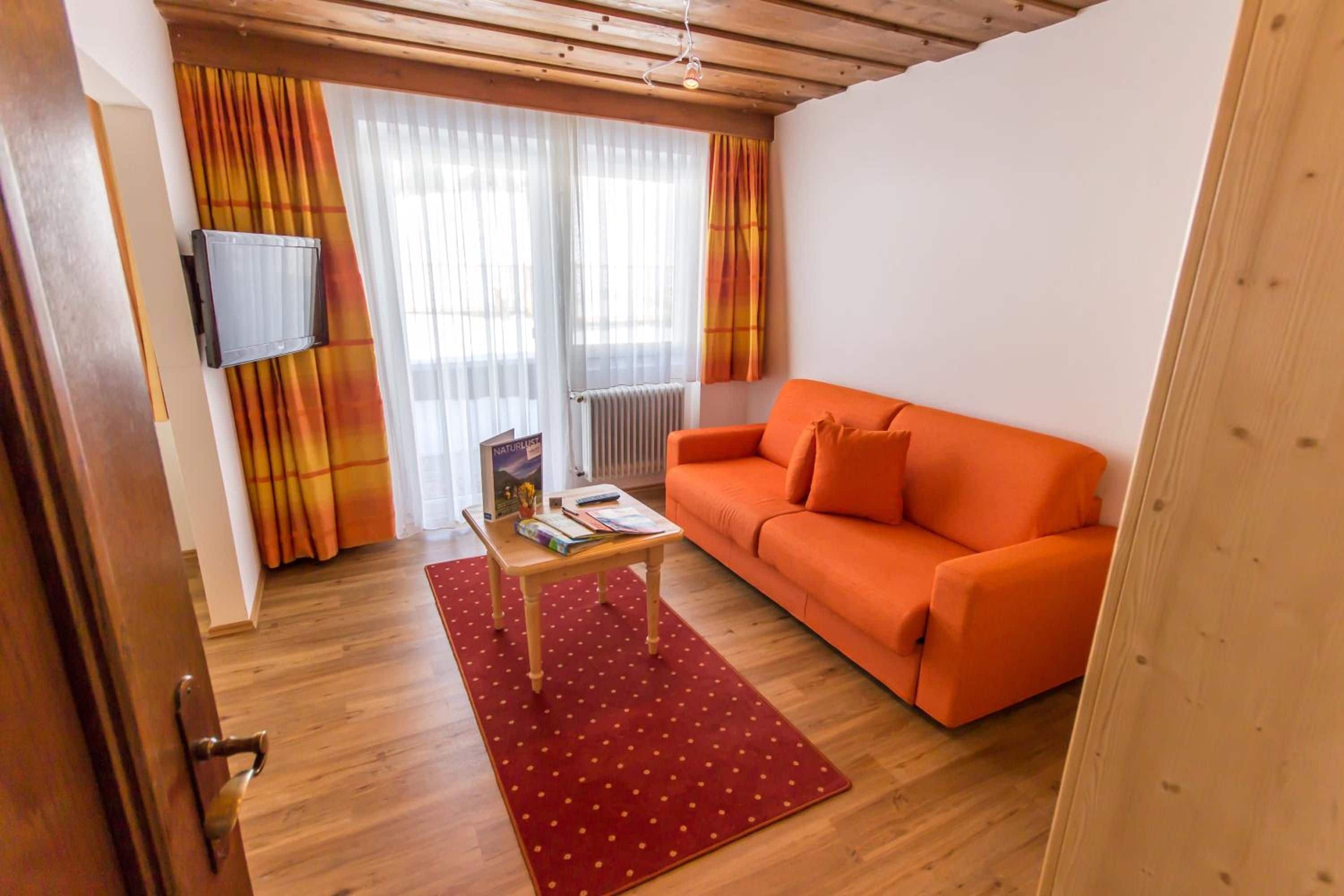 A cozy living room with a wooden ceiling, a red polka dot rug, an orange sofa, and a flat-screen TV mounted on the wall.