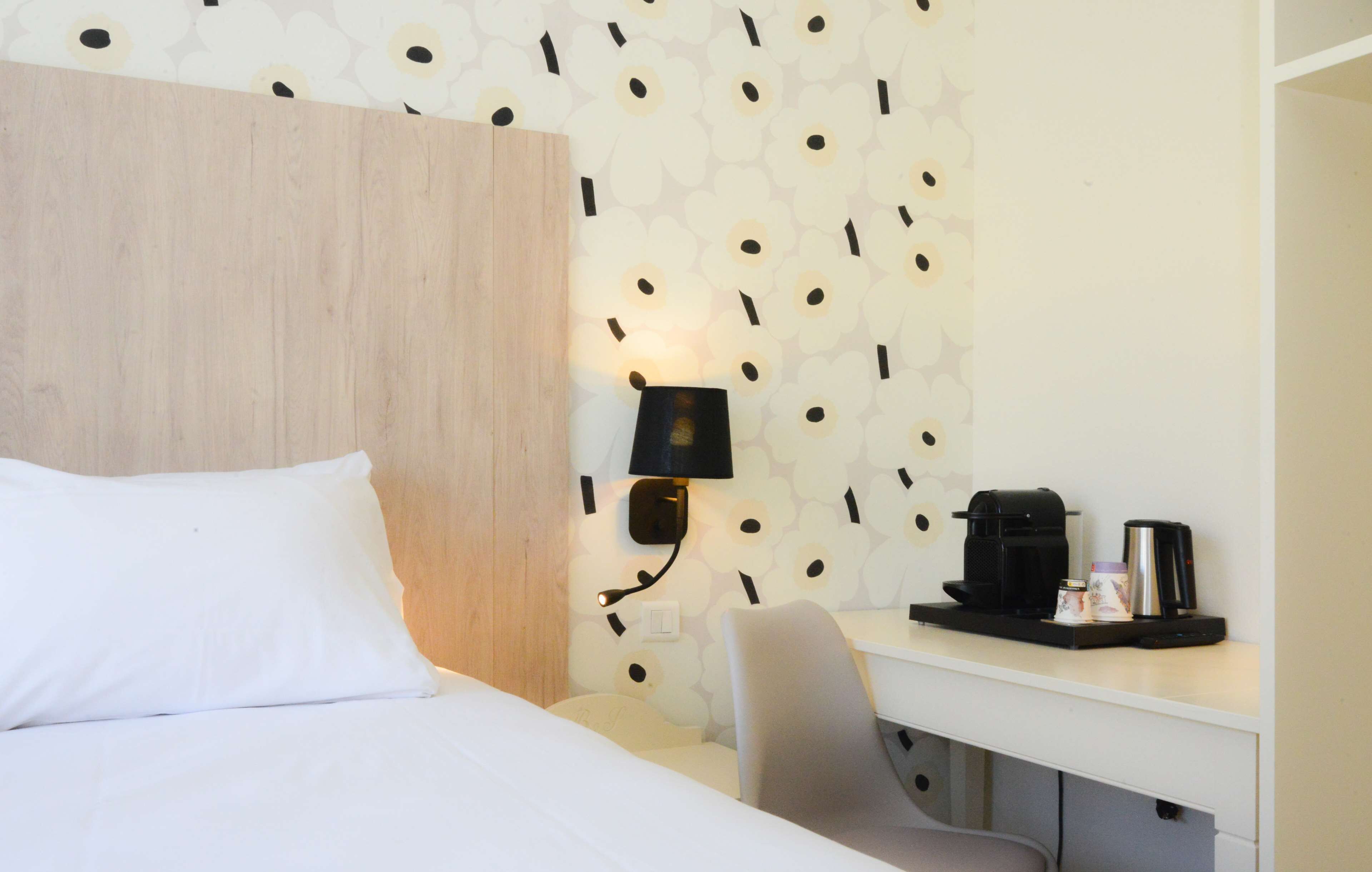 Hotelkamer met een bed, een bureau met lamp, een koffiezetapparaat en een wand met bloemenmotief.