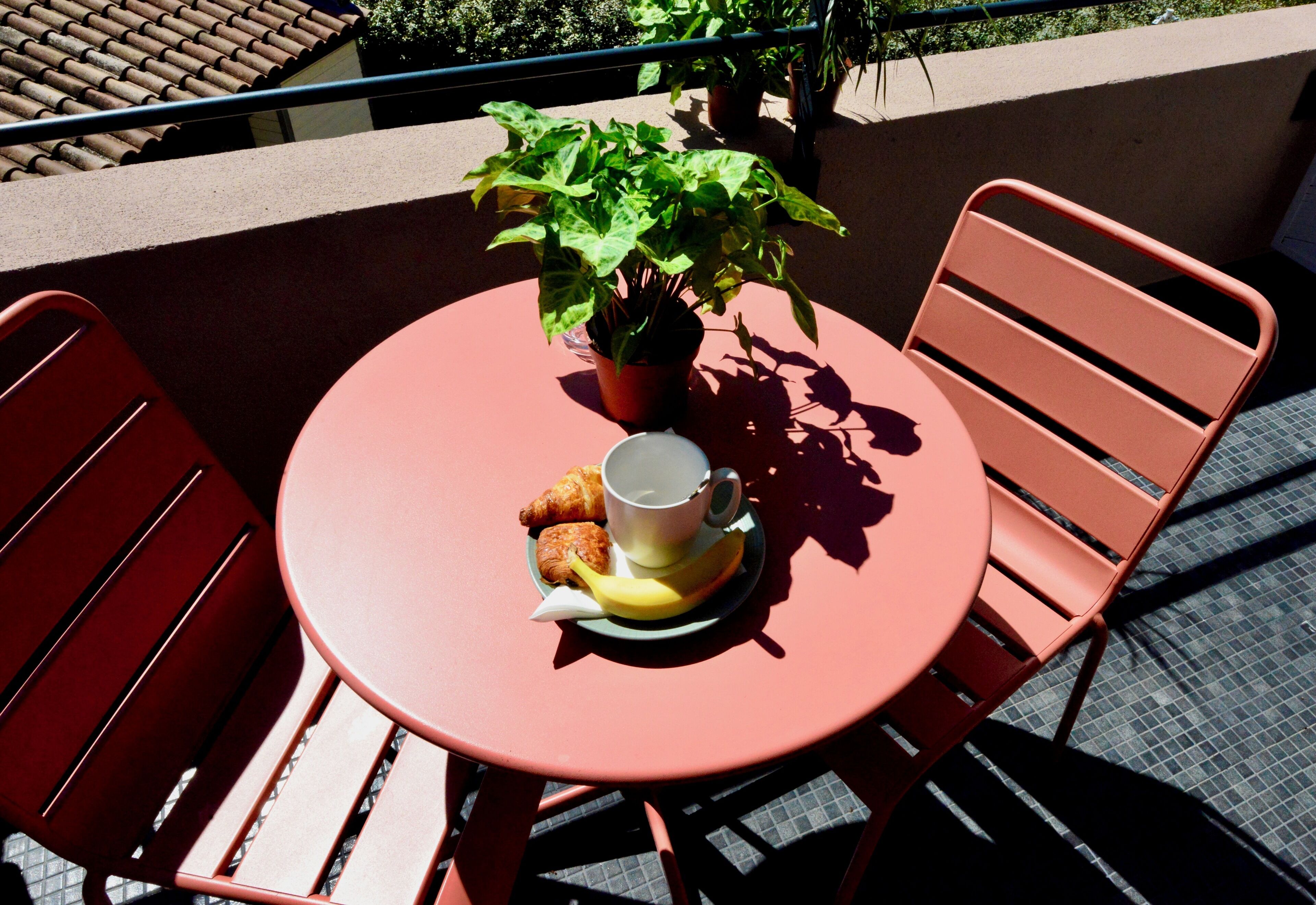 Una mesa redonda con una planta en maceta, un croissant, una taza y un plátano sobre un mantel rojo.