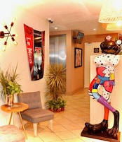 A lobby with a grey armchair, a potted plant, a wall-mounted light fixture, and a poster of a car.