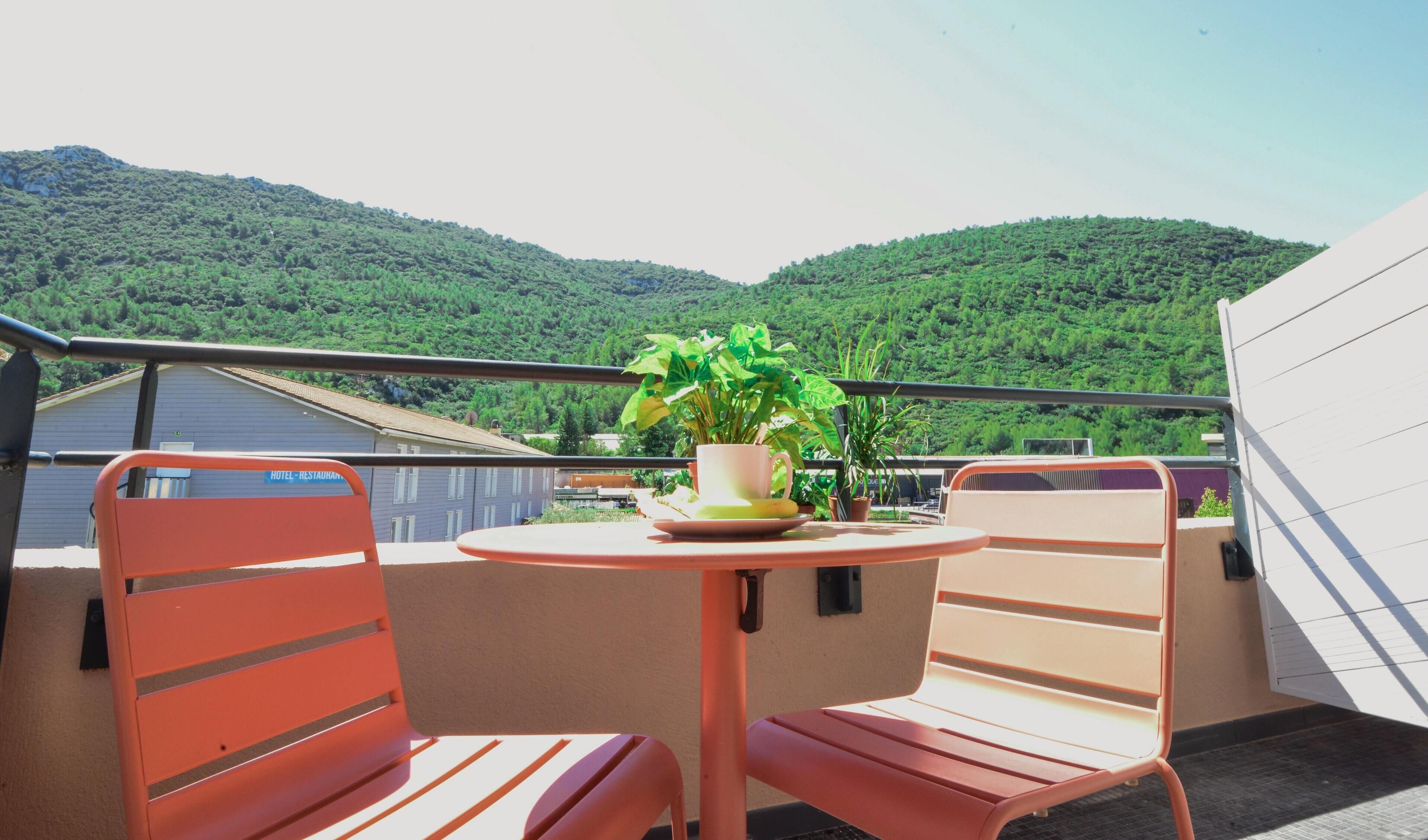 A balcony with two red chairs and a table, a potted plant, and a view of green hills.