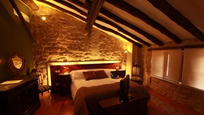Une chambre avec un mur en pierre, des poutres en bois, un lit avec des oreillers, un bureau, une chaise, une commode et une fenêtre avec des rideaux.