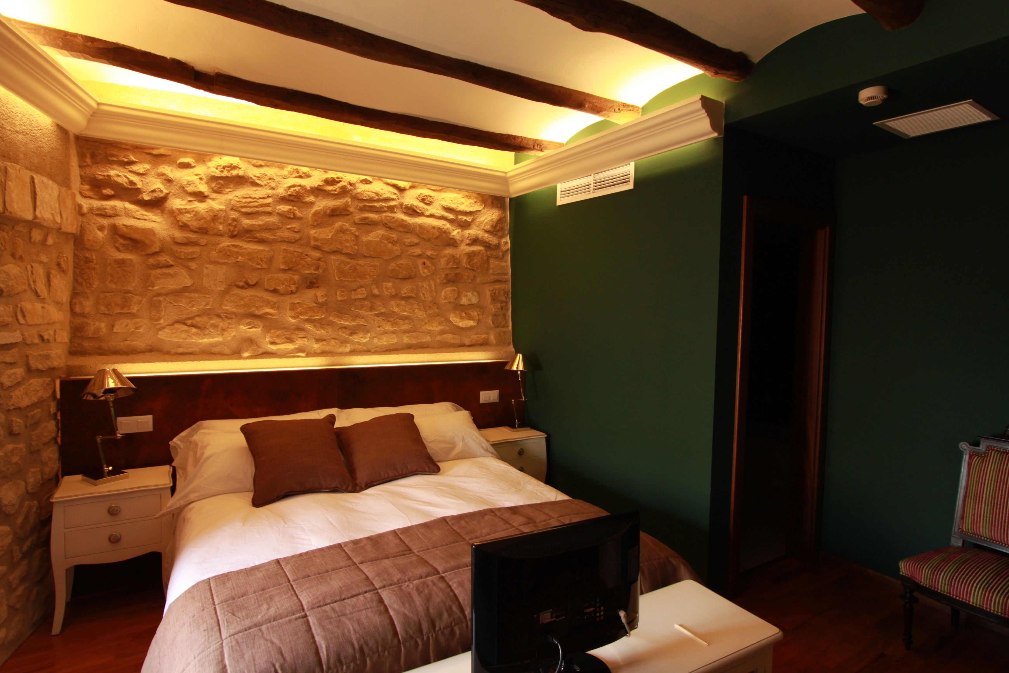 A bedroom with a stone wall, a wooden ceiling, a bed with a brown bedspread, a nightstand, a chair, and a desk with a computer.