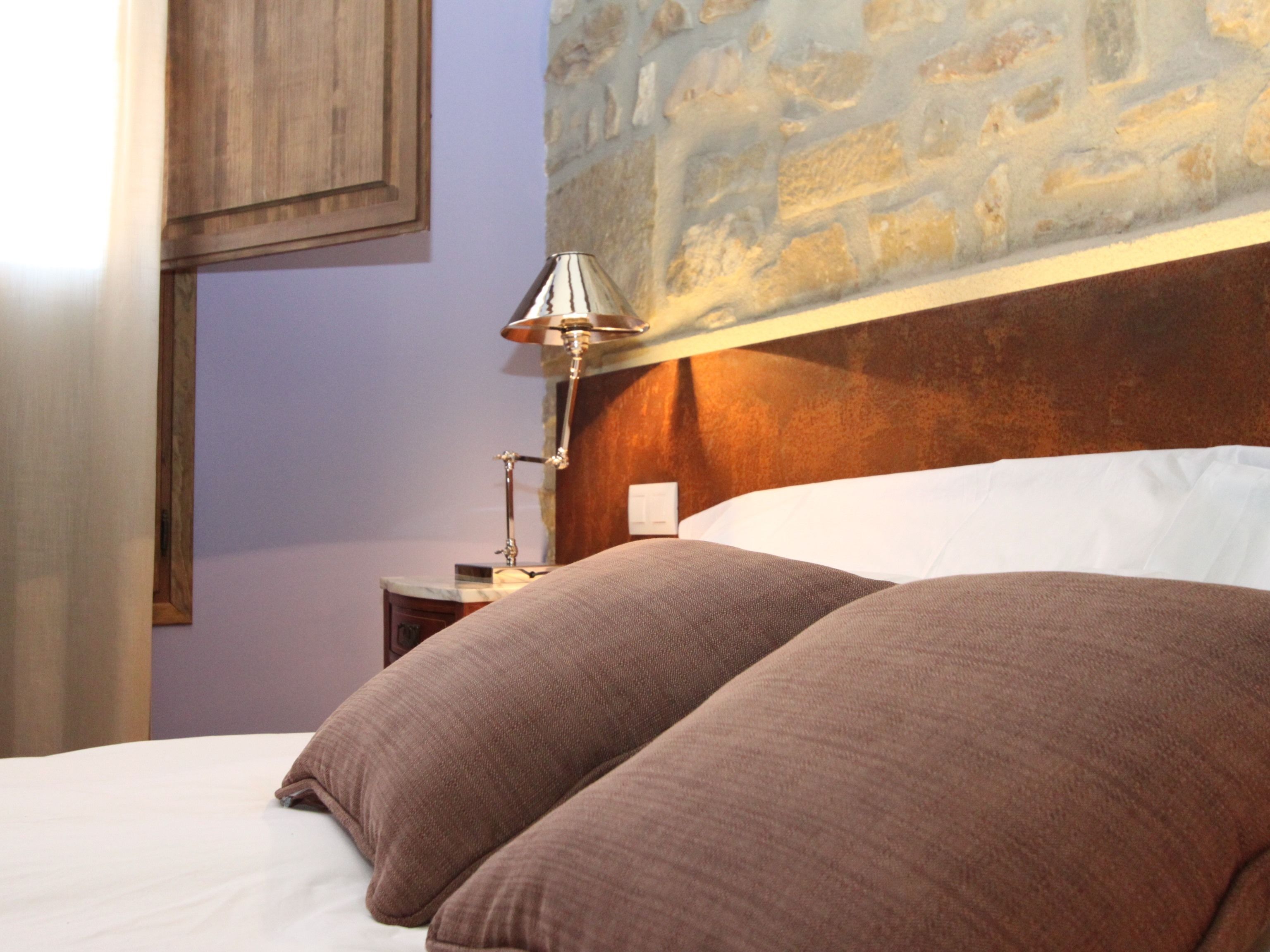 Una cama con dos almohadas, una mesita de noche con una lámpara y una pared de piedra.