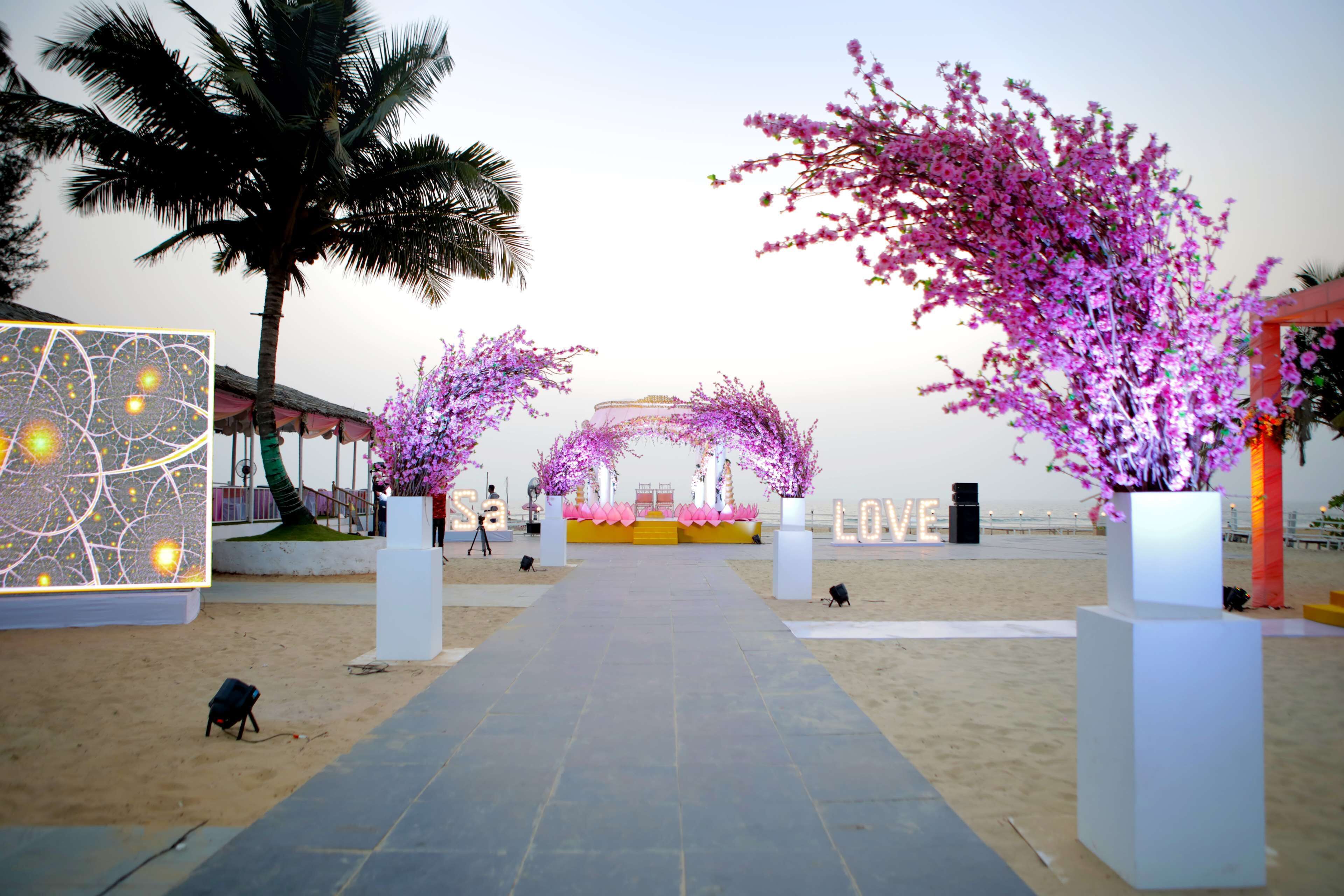 A beach walkway with a stage, palm trees, and pink flowering plants.