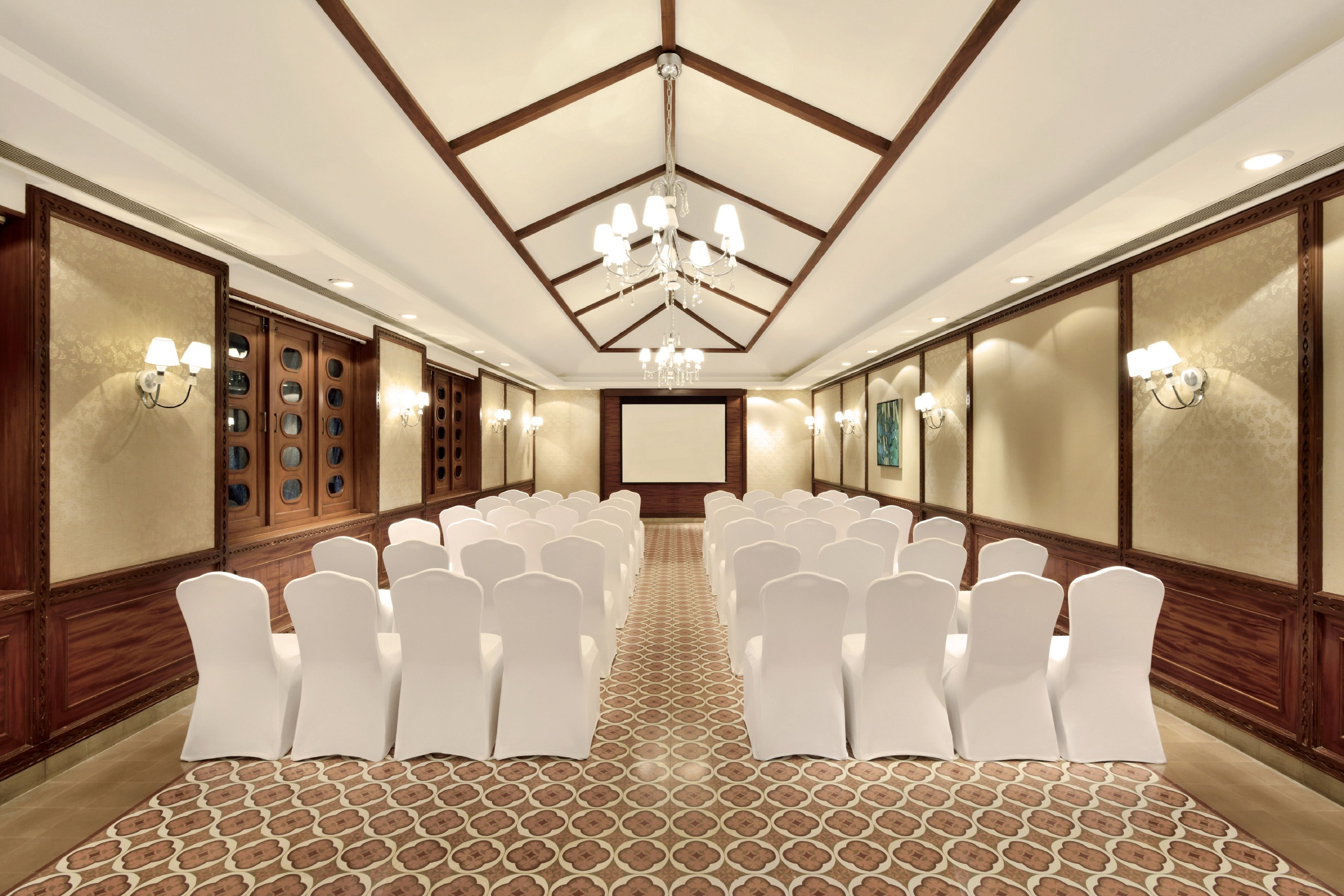 Un salón de conferencias con filas de sillas blancas, una alfombra estampada, paneles de madera y apliques.