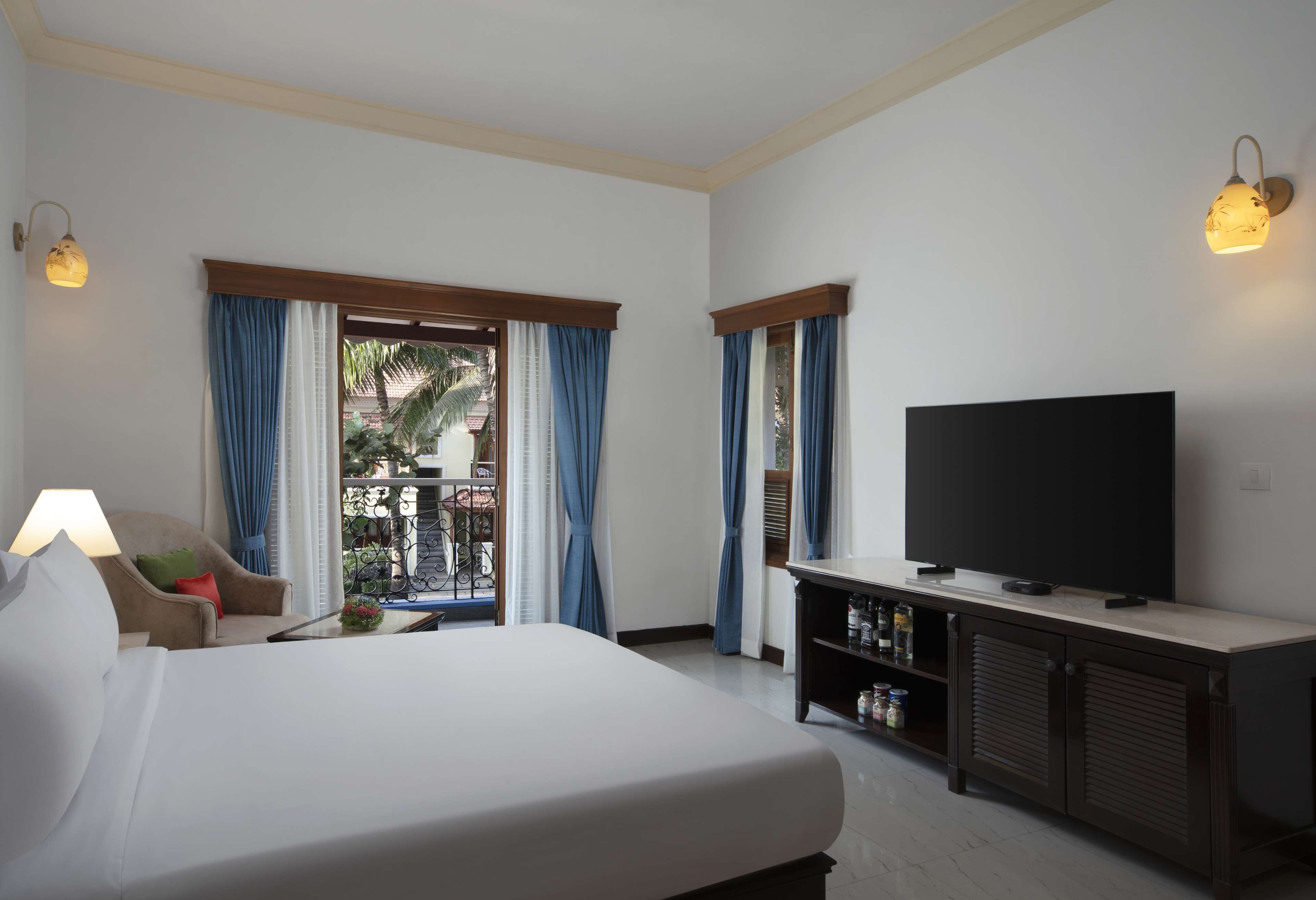 Habitació d'hotel amb un llit, una televisió sobre un moble, una cadira amb una làmpada i un balcó amb cortines.