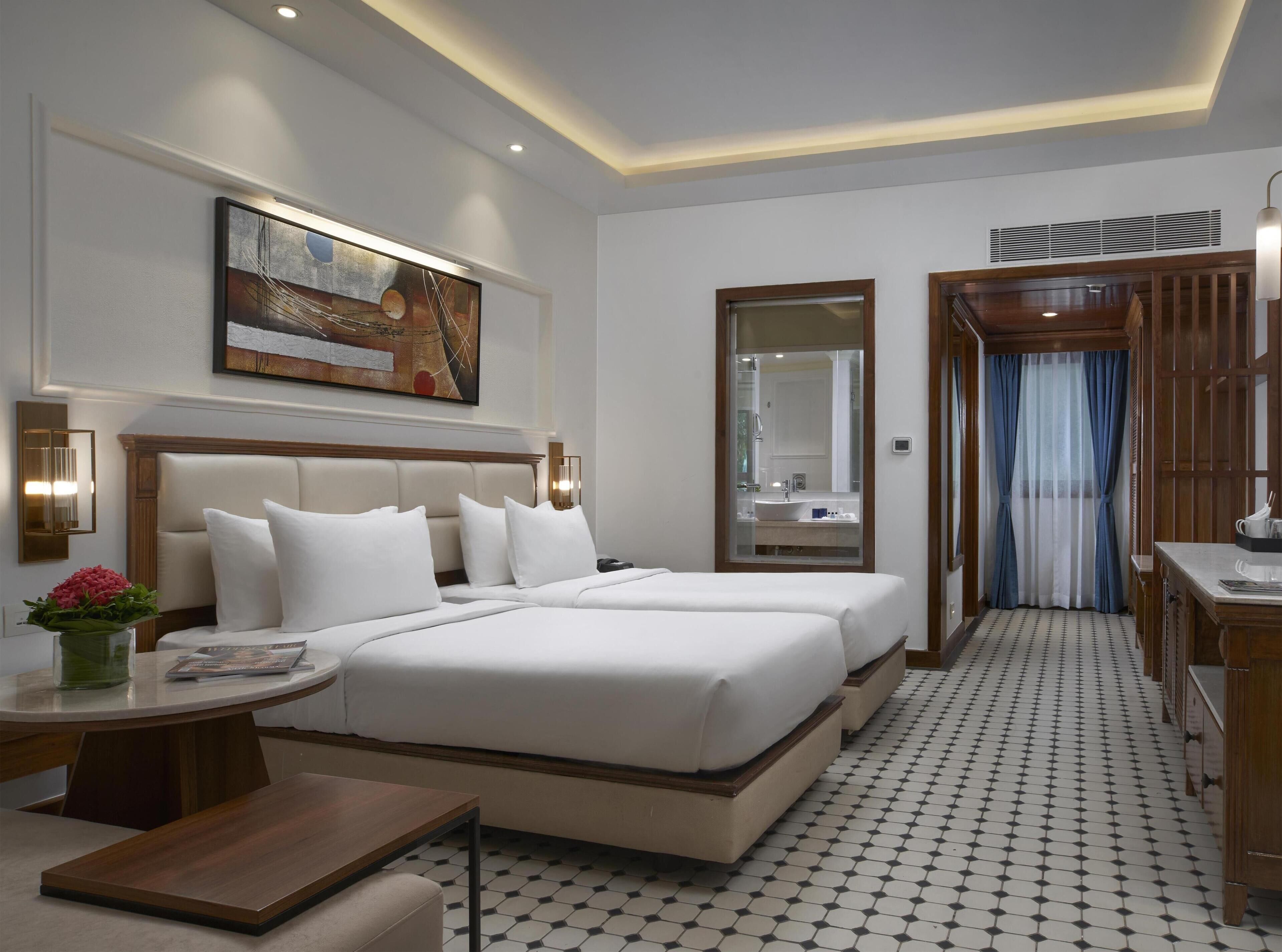 A hotel room with a large bed, a desk, a chair, a small table, a vase with flowers, a framed picture on the wall, a mirror, and a patterned floor.