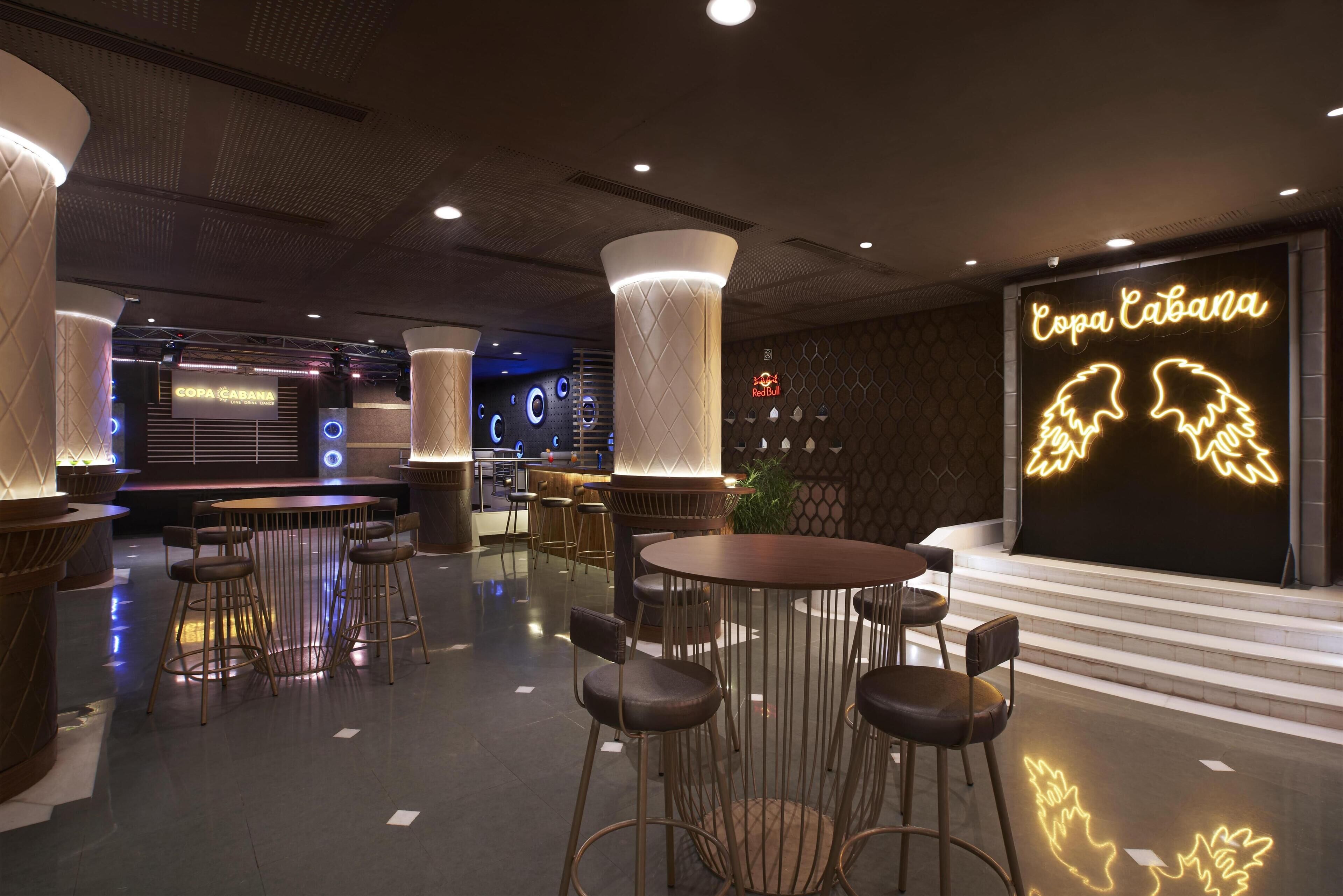 Ein modernes Interieur mit runden Tischen und Barhocker, einer Neonreklame mit der Aufschrift "Copa Cabana" und einer Sitzecke mit gemustertem Wanddesign und Blumentöpfen.