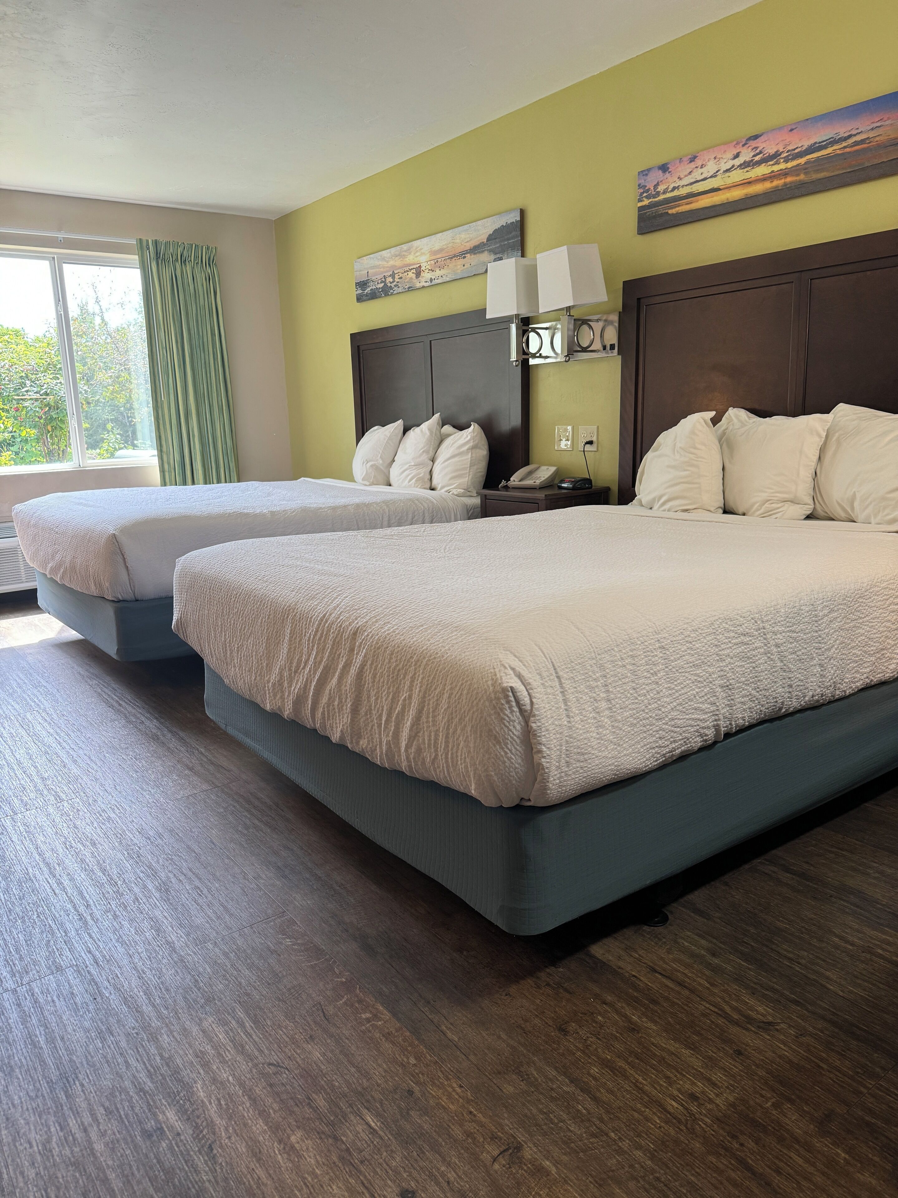 A hotel room with two beds, a large window with curtains, a painting on the wall, and a nightstand with a lamp.