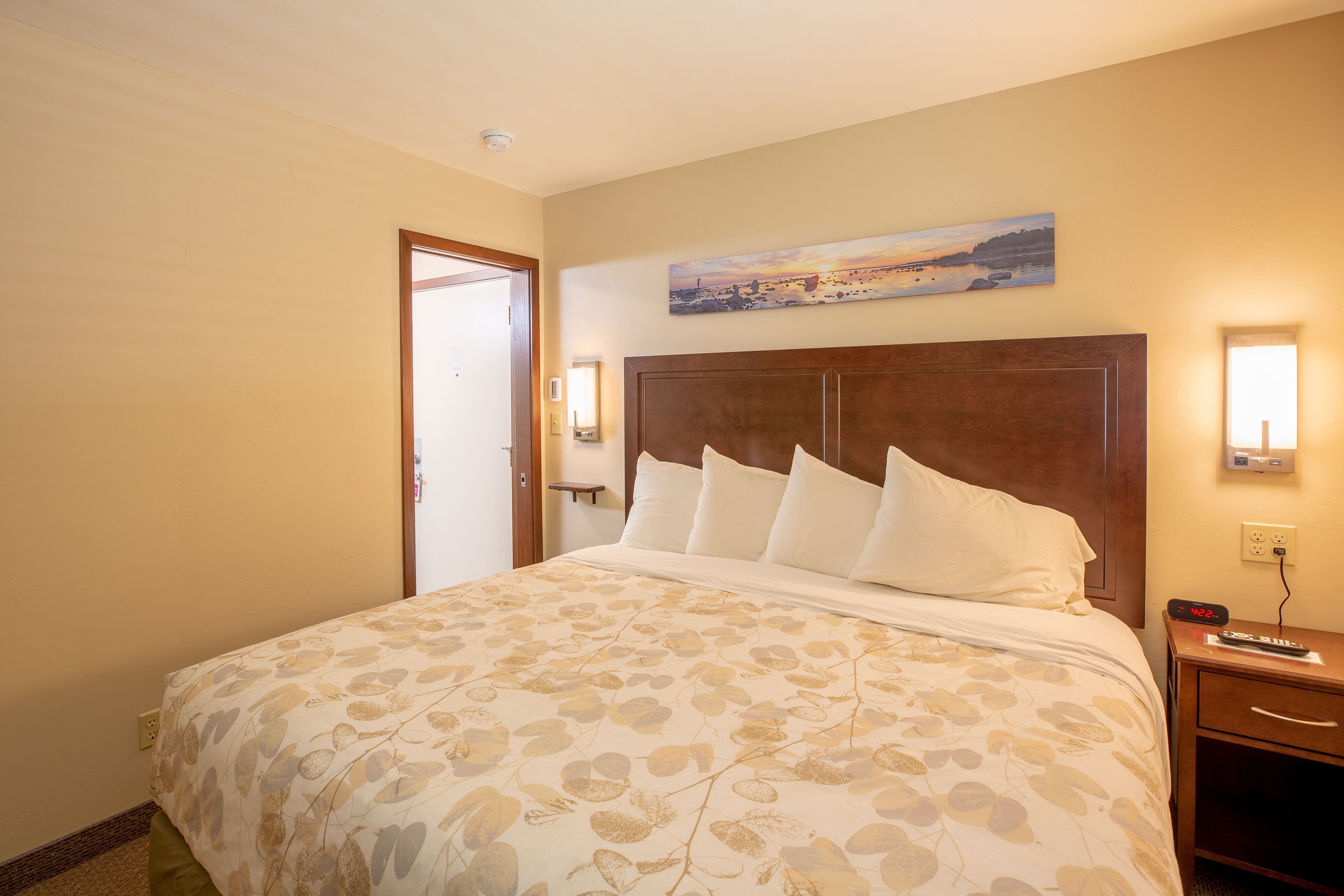 Une chambre d’hôtel avec un grand lit, une tête de lit en bois, un tableau au mur et une table de chevet avec une horloge.