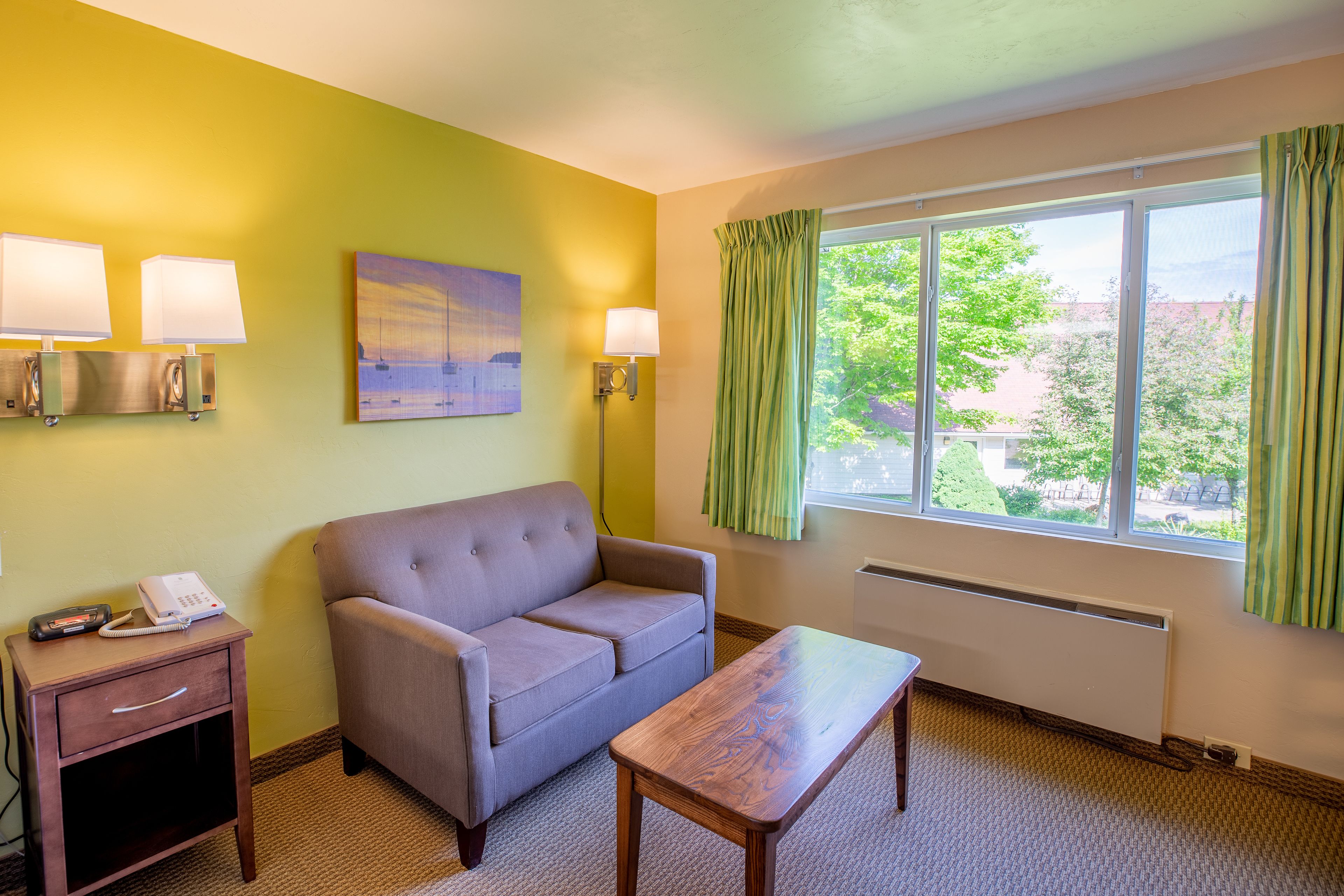 A hotel room with a sofa, a small table, a bedside table with a phone, two wall lamps, a painting, and a window with curtains.