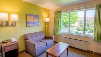 A hotel room with a sofa, a small table, a bedside table with a phone, two wall lamps, a painting, and a window with curtains.