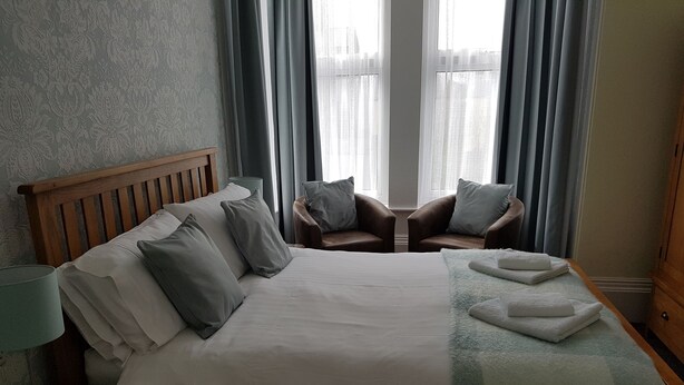 A neatly made bed with pillows, a bedside lamp, and a checkered throw blanket. There are two armchairs with cushions and a window with curtains. - The Tennyson (Bridlington)