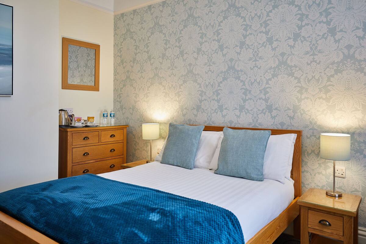A neatly made bed with a blue blanket, two bedside lamps, a wooden nightstand, a wooden dresser, and a framed picture on the wall.