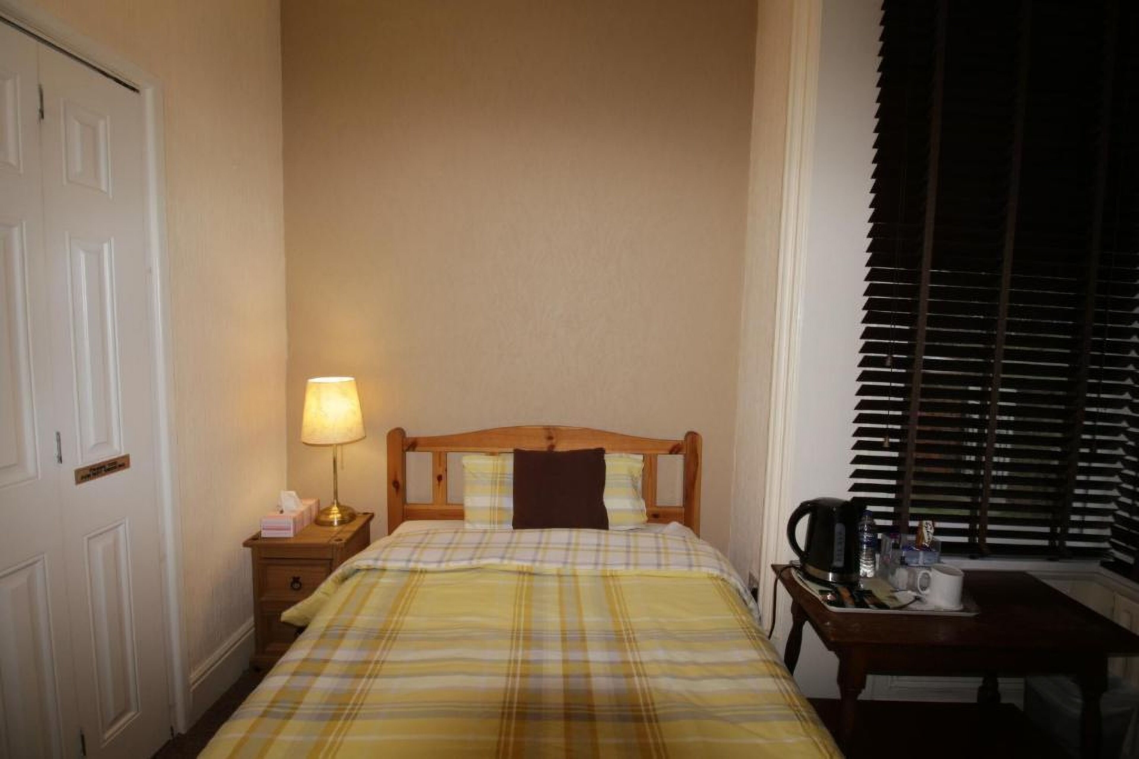 A hotel room with a bed, a bedside table with a lamp, a coffee maker, and a window with blinds.