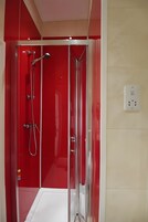Cabina de ducha con paredes de vidrio, grifería cromada y revestimiento rojo.