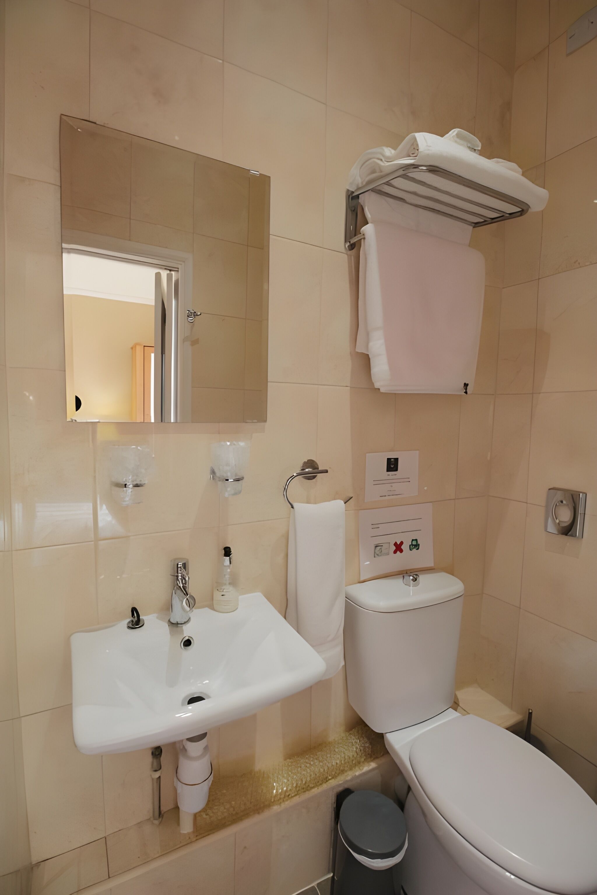 A bathroom with a white sink, mirror, and toilet. There is a towel rack above the toilet and a trash bin next to it.