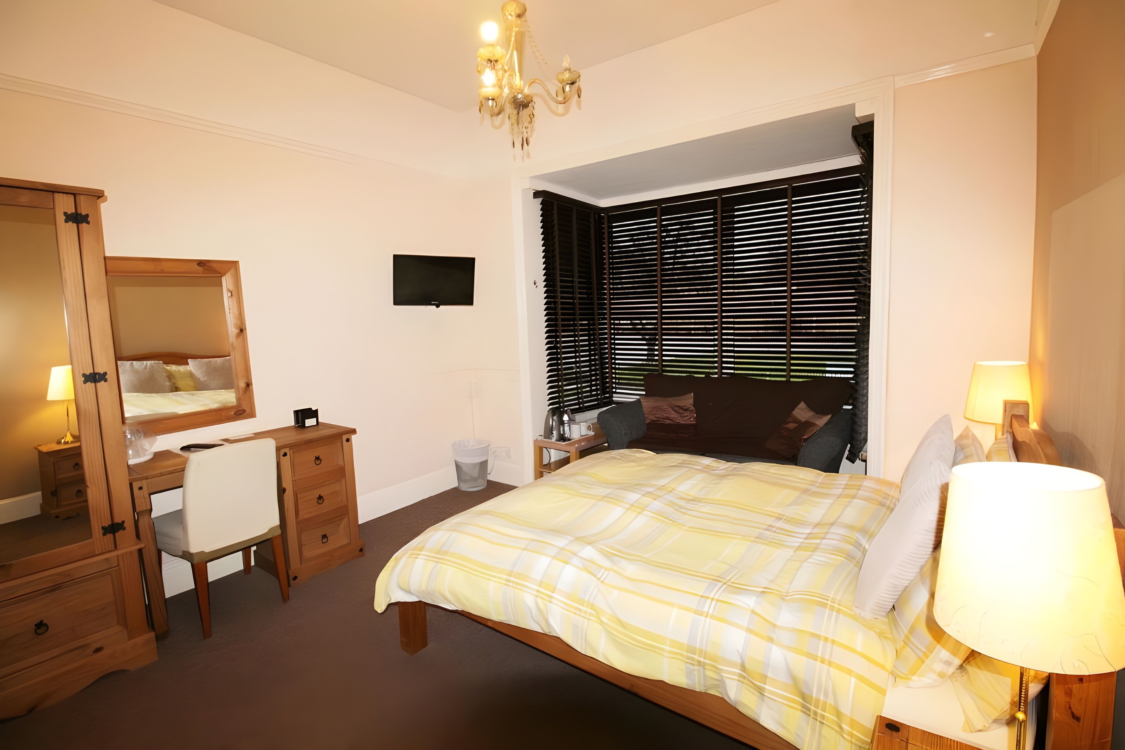A bedroom with a bed, a desk, a chair, a dresser, a mirror, a TV, and a window with blinds.