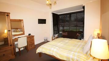 A bedroom with a bed, a desk, a chair, a dresser, a mirror, a TV, and a window with blinds.