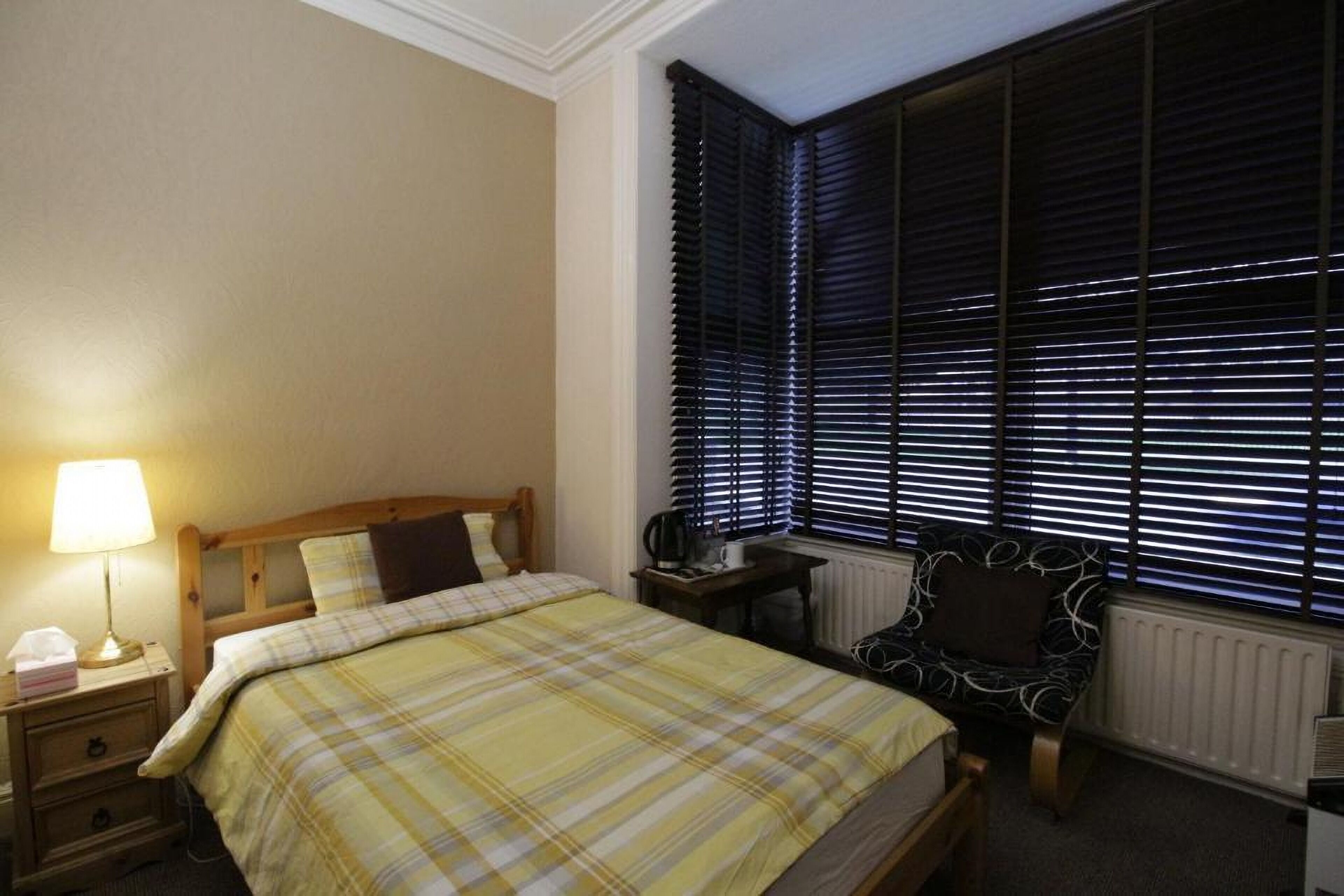Une chambre à coucher avec un lit, une table de chevet avec une lampe, une chaise et une fenêtre avec des stores.
