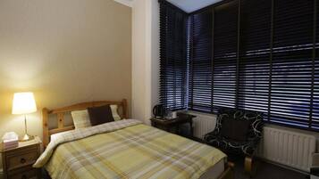 Un dormitorio con cama, una mesita de noche con lámpara, una silla y una ventana con persianas.