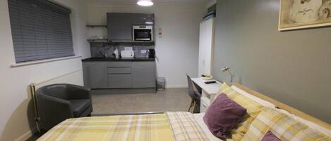 A compact hotel room with a bed, a desk, a chair, and a kitchenette.