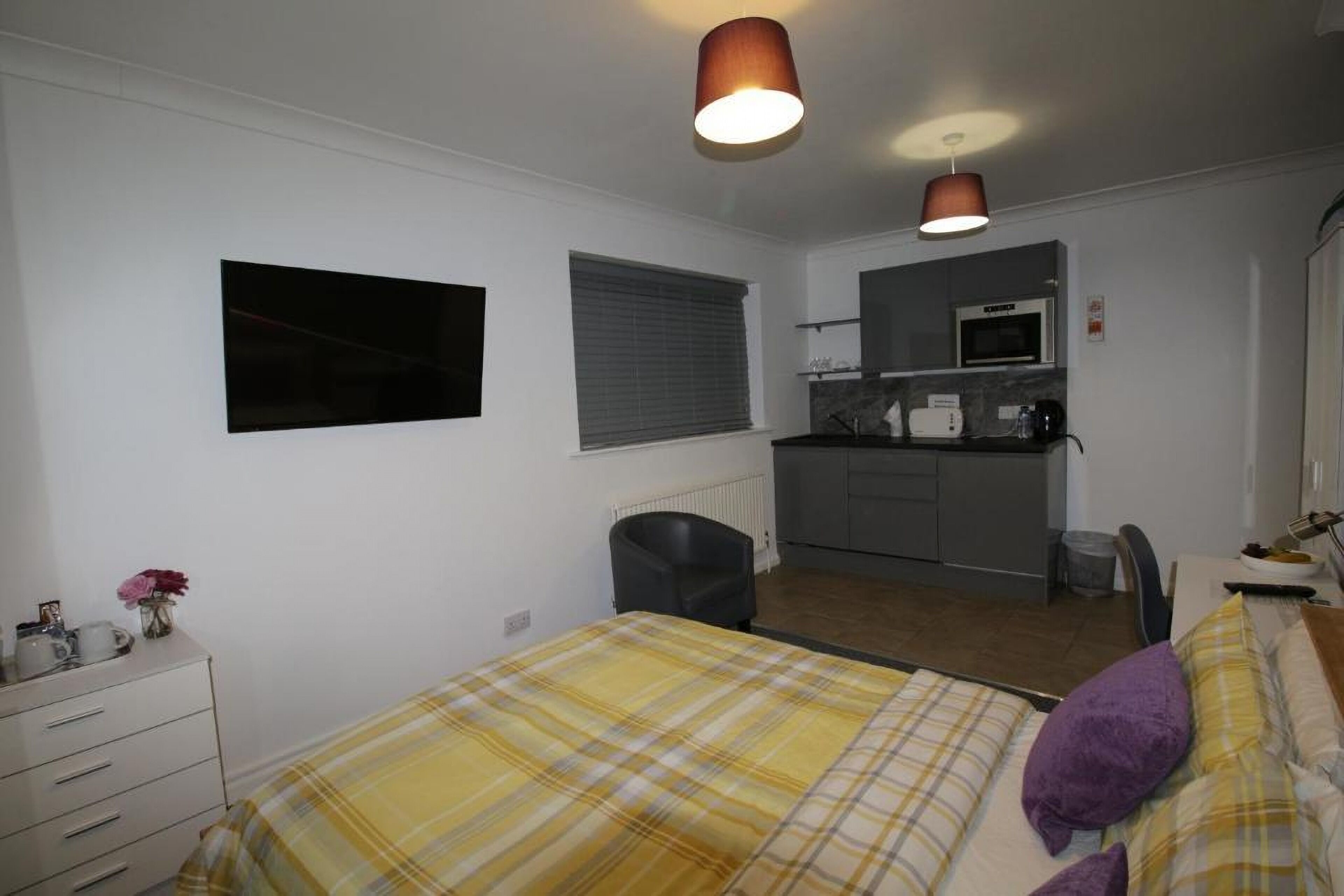 Ein Schlafzimmer mit Bett, Schreibtisch, Stuhl, einer kleinen Küchenzeile und einem Fenster mit Jalousien.