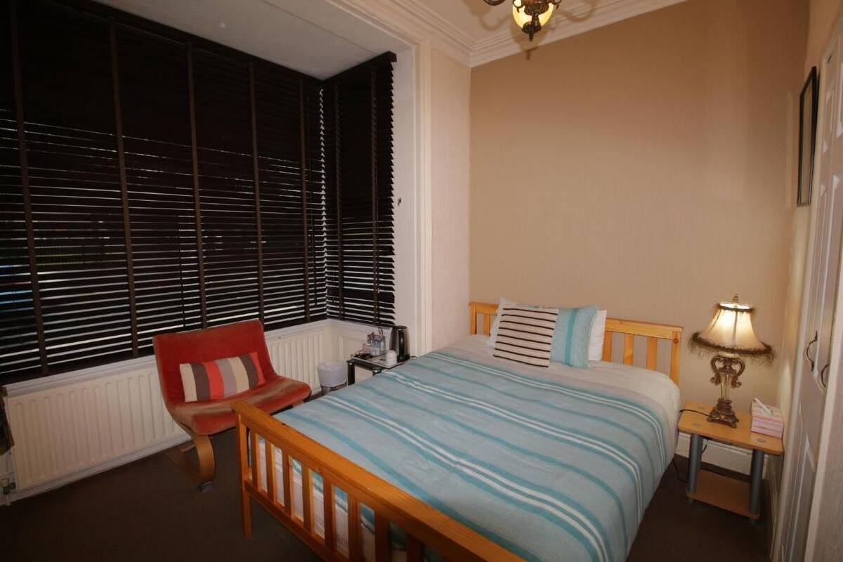 Quarto com cama, cadeira, mesa de cabeceira, abajur e janela com persianas.