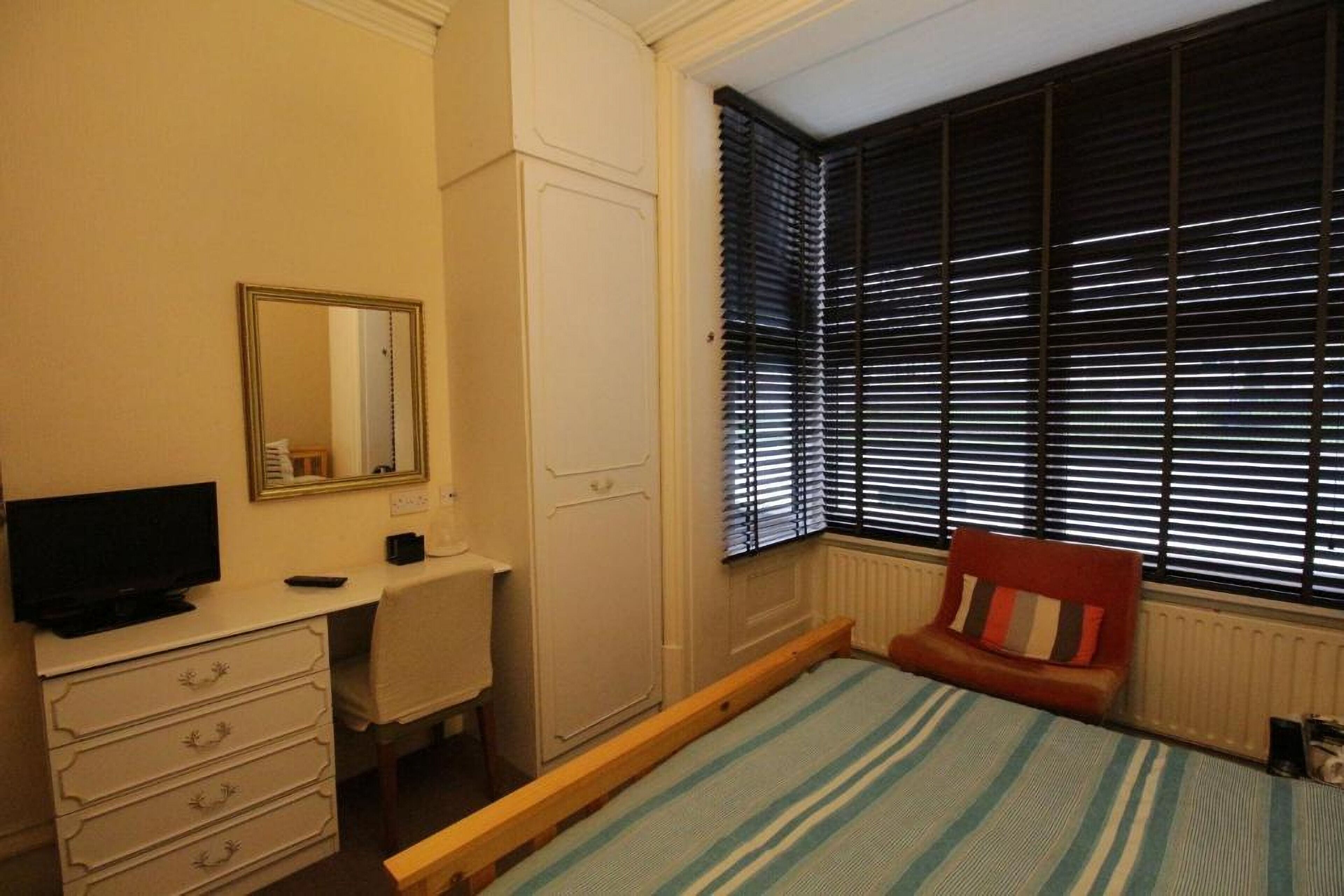 Ein Schlafzimmer mit einem Bett, einem Schreibtisch, einem Fernseher, einem Kleiderschrank und einem Fenster mit Jalousien.